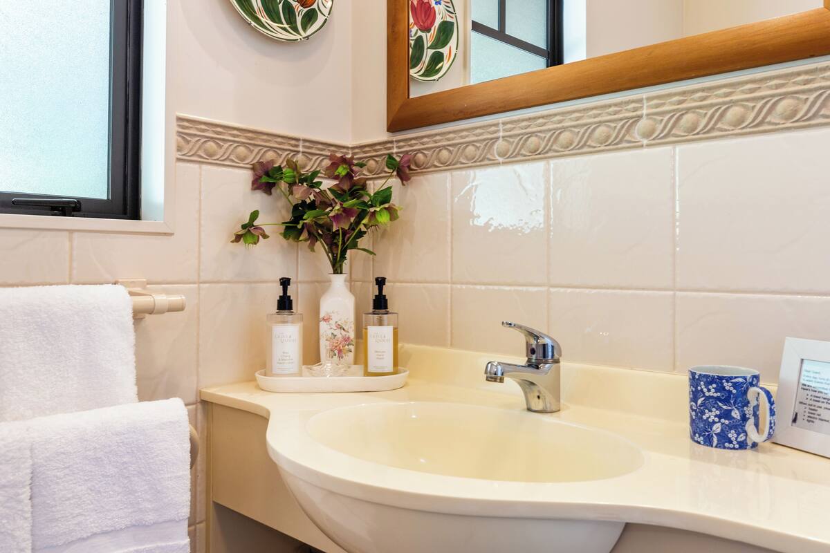 A bathroom with a sink, a mirror, a window, and a vase with flowers.