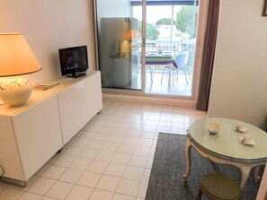 A modern hotel room with a TV on a white cabinet, a glass table with a chair, and a view of the outdoors through large windows.