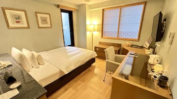 A hotel room with a bed, a desk with a computer, a chair, a lamp, a TV, and a window with blinds.