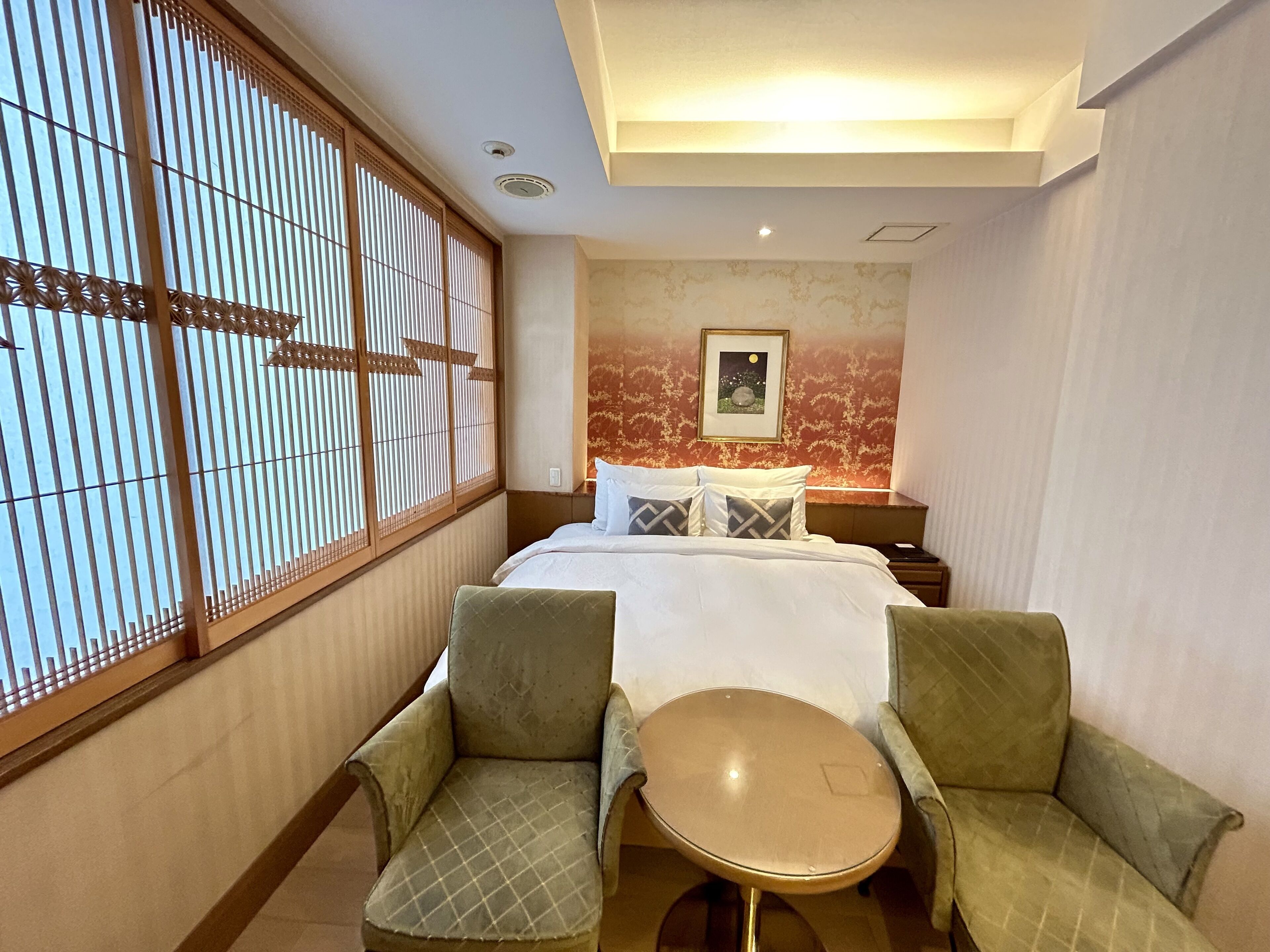 Una habitación de hotel con cama, dos sillones, una mesita redonda, una mesita de noche y una ventana con persianas verticales.