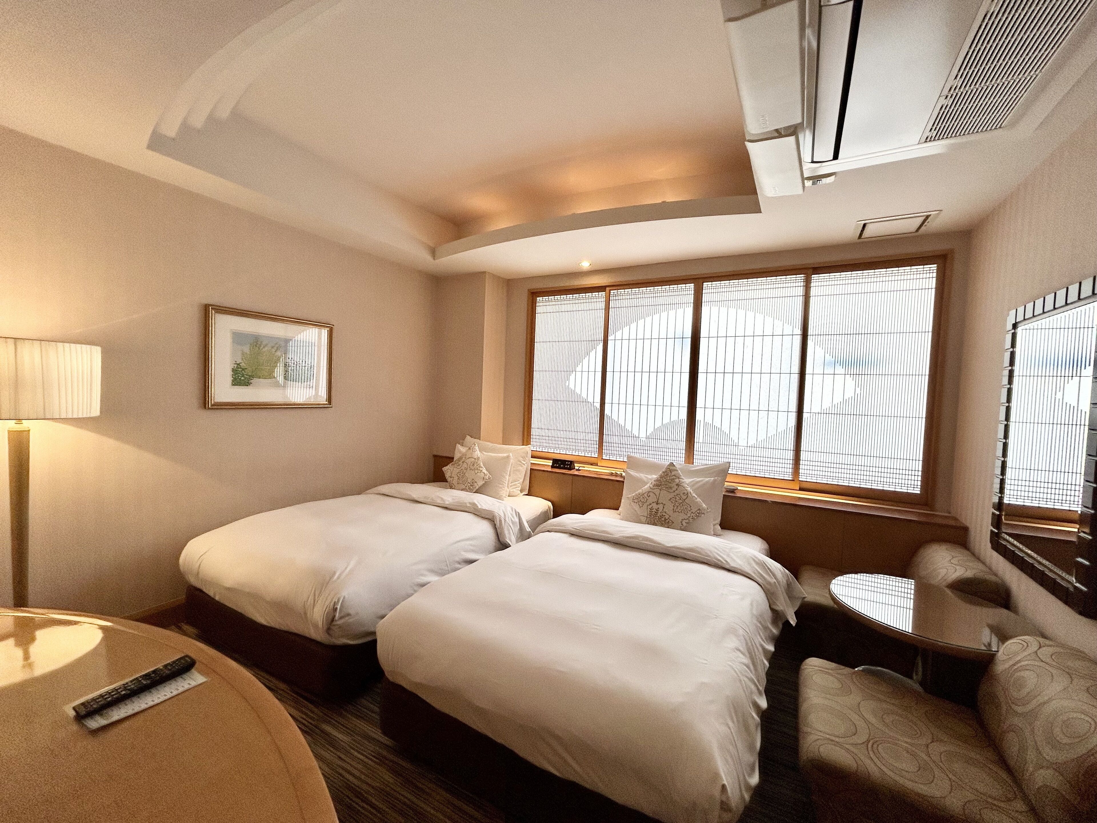 A hotel room with two beds, a bedside table, a chair, a lamp, and a window with blinds.