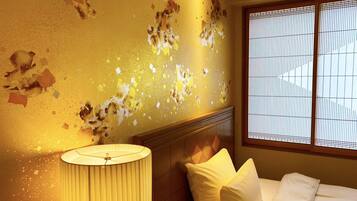A hotel room with peeling wall paint, a bed with white bedding, a bedside lamp, and a window with blinds.