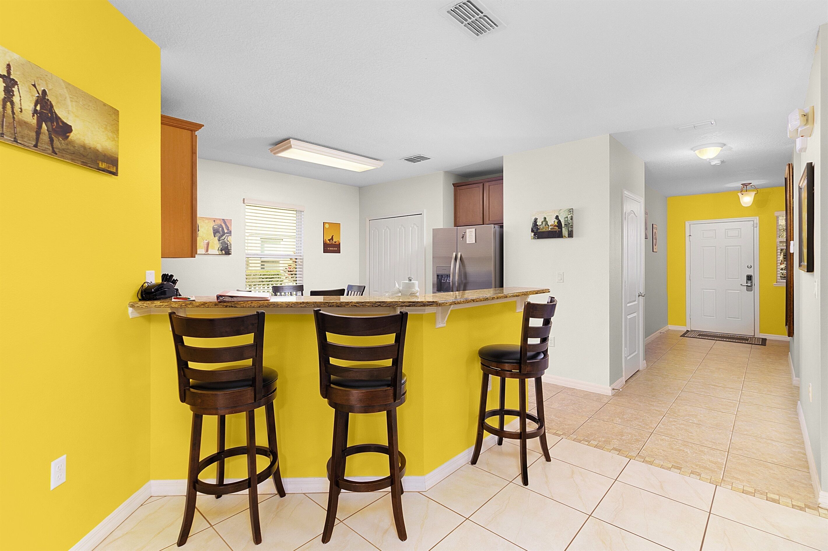 A kitchen with a central island, three bar stools, a refrigerator, and a window with blinds.