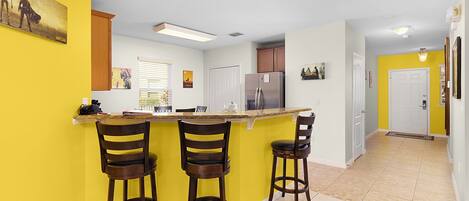 A kitchen with a central island, three bar stools, a refrigerator, and a window with blinds.