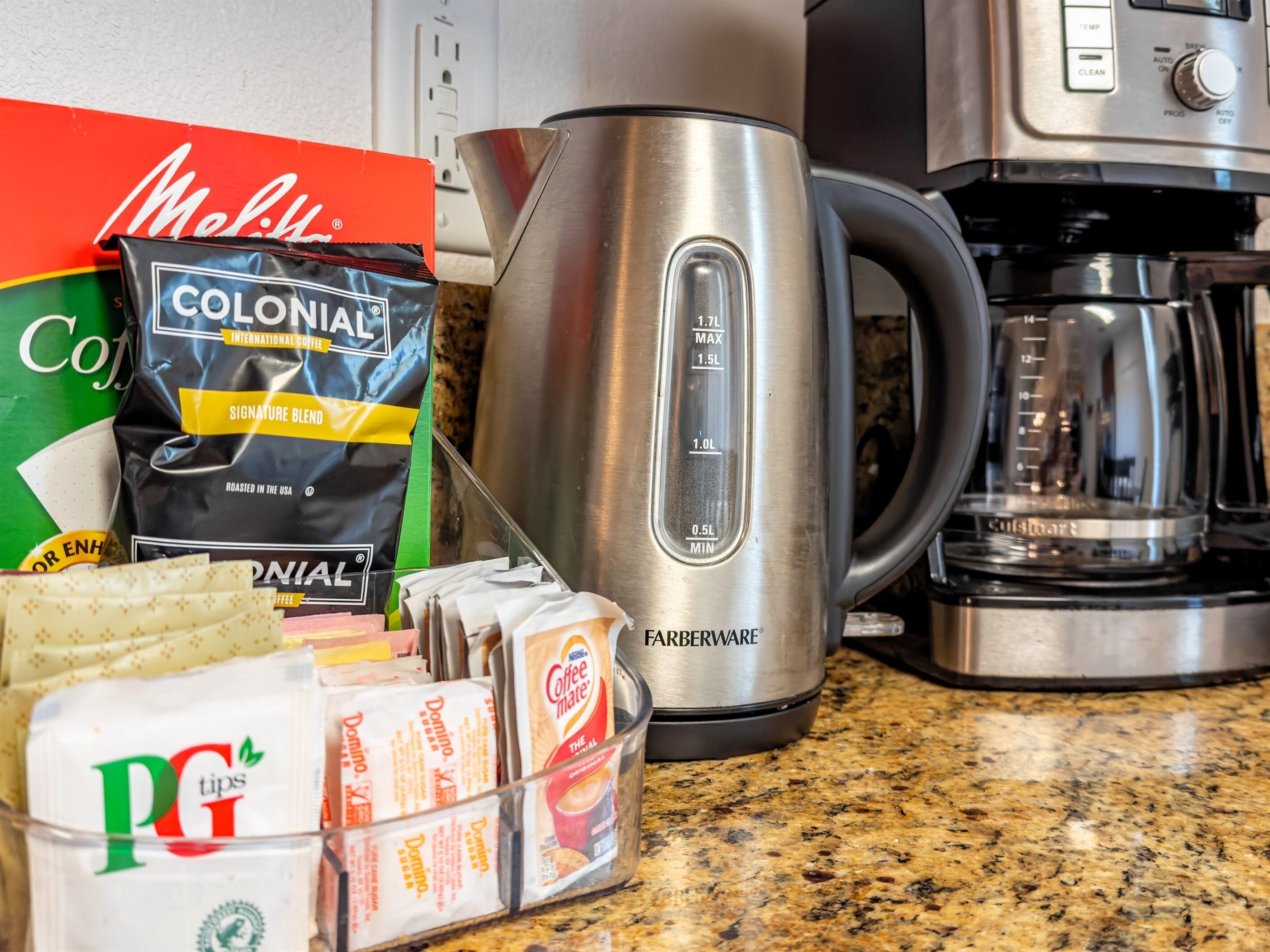 A kitchen counter with a coffee maker, a kettle, and various coffee pods and packets.