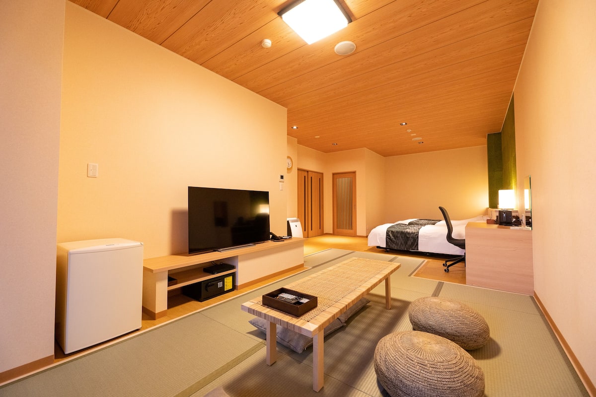 Ein modernes Hotelzimmer mit Holzboden, einem Flachbildfernseher, einem Bett mit weißer Bettdecke, einem Schreibtisch mit Computer und Tatami-Böden.
