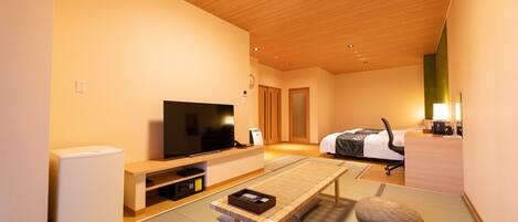 A modern hotel room with a wooden ceiling, a flat-screen TV, a bed with a white comforter, a desk with a computer, and tatami flooring.