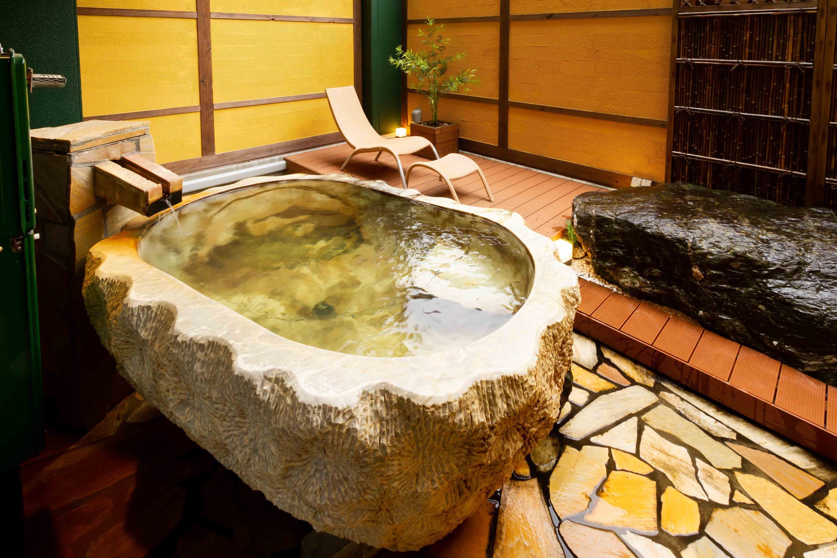 A stone hot tub with a wooden bench, a potted plant, and a tiled floor.