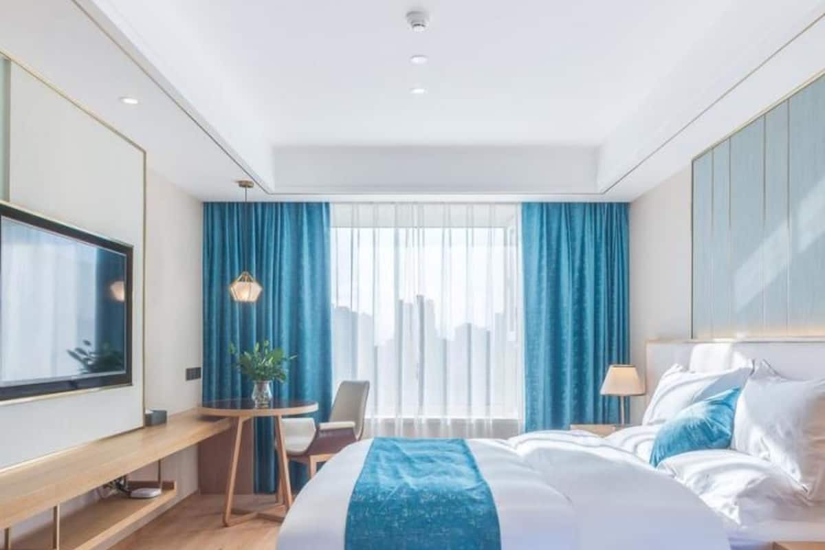 A modern hotel room with a large bed, a desk, a chair, a TV, and a window with curtains.
