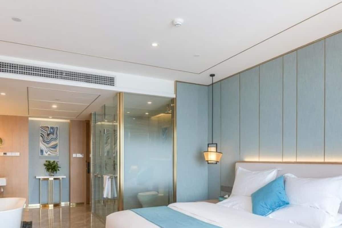 A modern hotel room with a large bed, a bedside table, a wall-mounted lamp, and a glass shower enclosure.