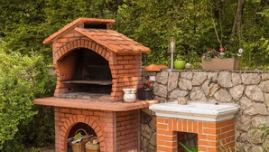 Ein Pizzaofen aus Backstein im Freien, mit einer Steinwand und Grünpflanzen im Hintergrund.