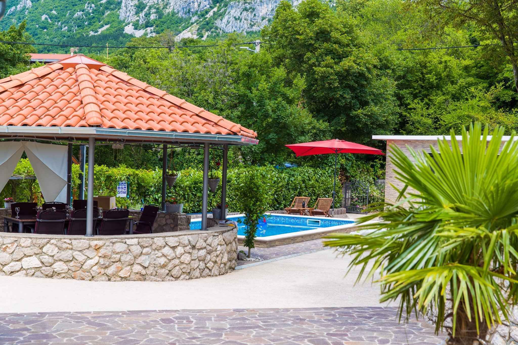 A covered outdoor seating area with a pool, surrounded by greenery and a mountain backdrop.