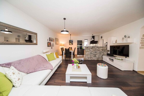 Una sala de estar moderna con un sofá blanco, una mesa de centro con objetos decorativos, un televisor y una cocina al fondo.