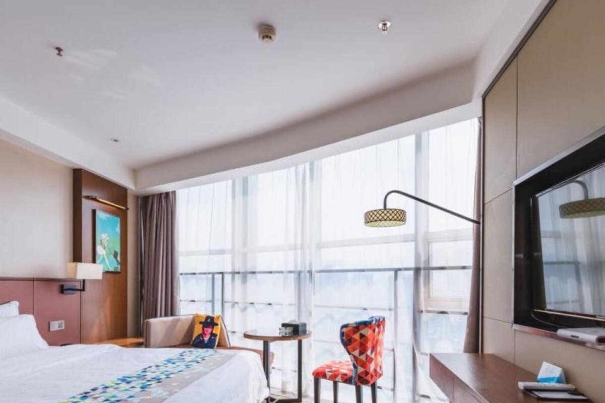 A hotel room with a bed, a desk, a chair, a TV, and large windows.