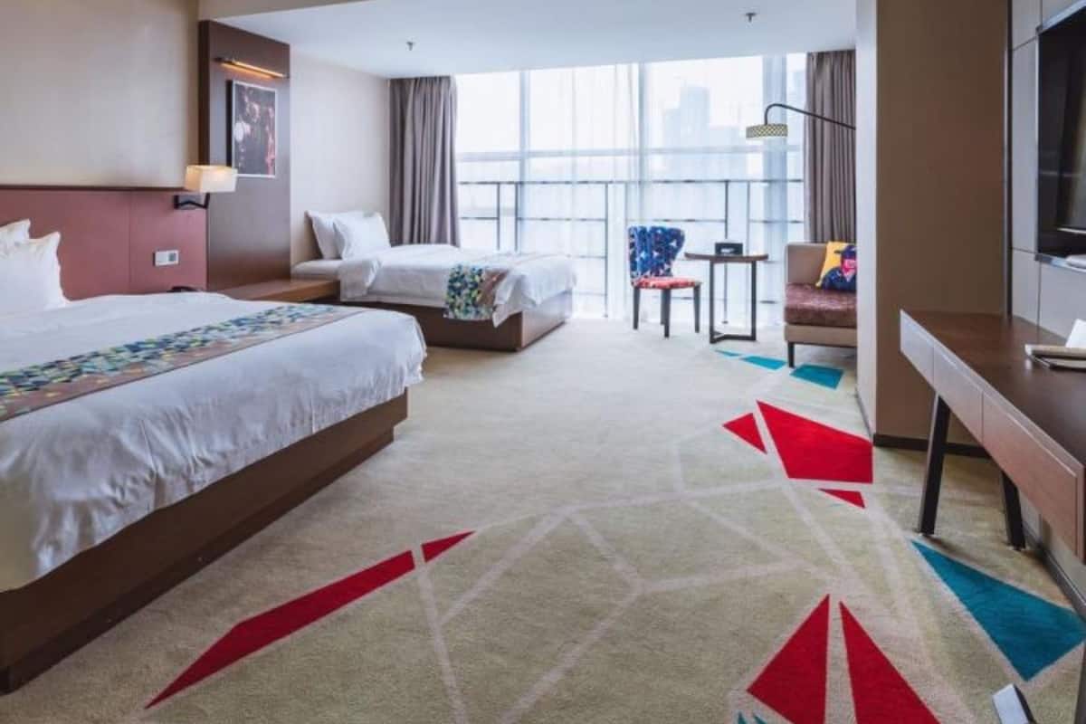 A hotel room with two beds, a desk, a chair, and a large window offering a city view.