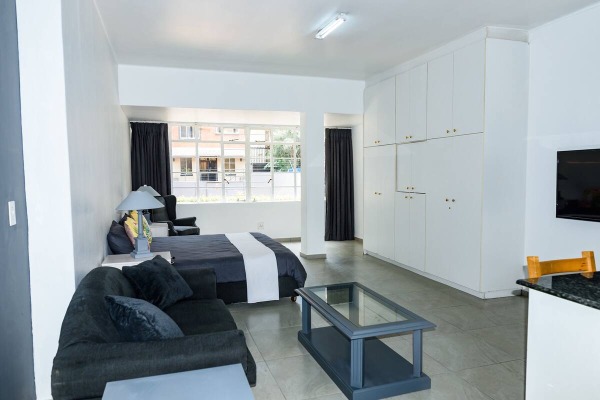 A modern, well-lit room with a bed, sofa, and a glass coffee table. There is a large window with curtains, a TV mounted on the wall, and a wardrobe with multiple doors.