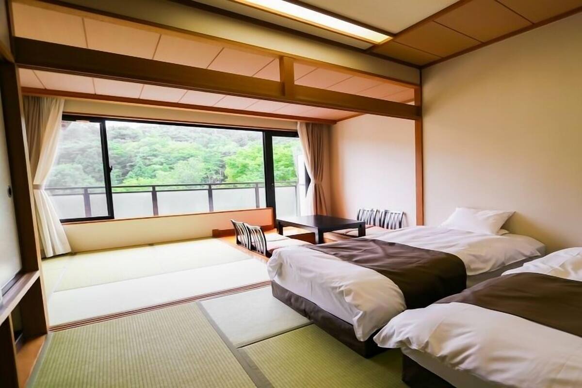A hotel room with two beds, a table, and a view of greenery outside.