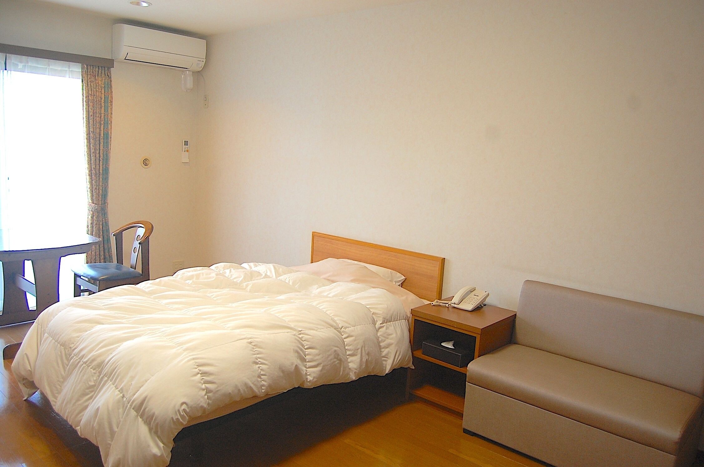 A hotel room with a bed, a bedside table with a phone, a sofa, and a window with curtains.