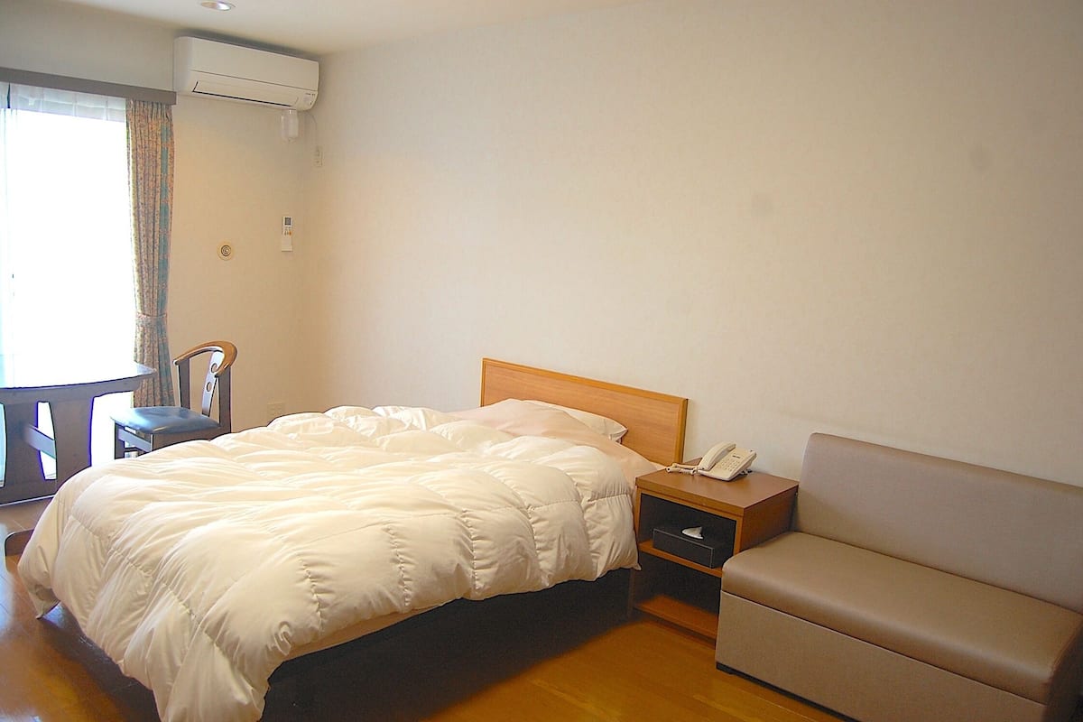 A hotel room with a bed, a bedside table with a phone, a sofa, and a window with curtains.