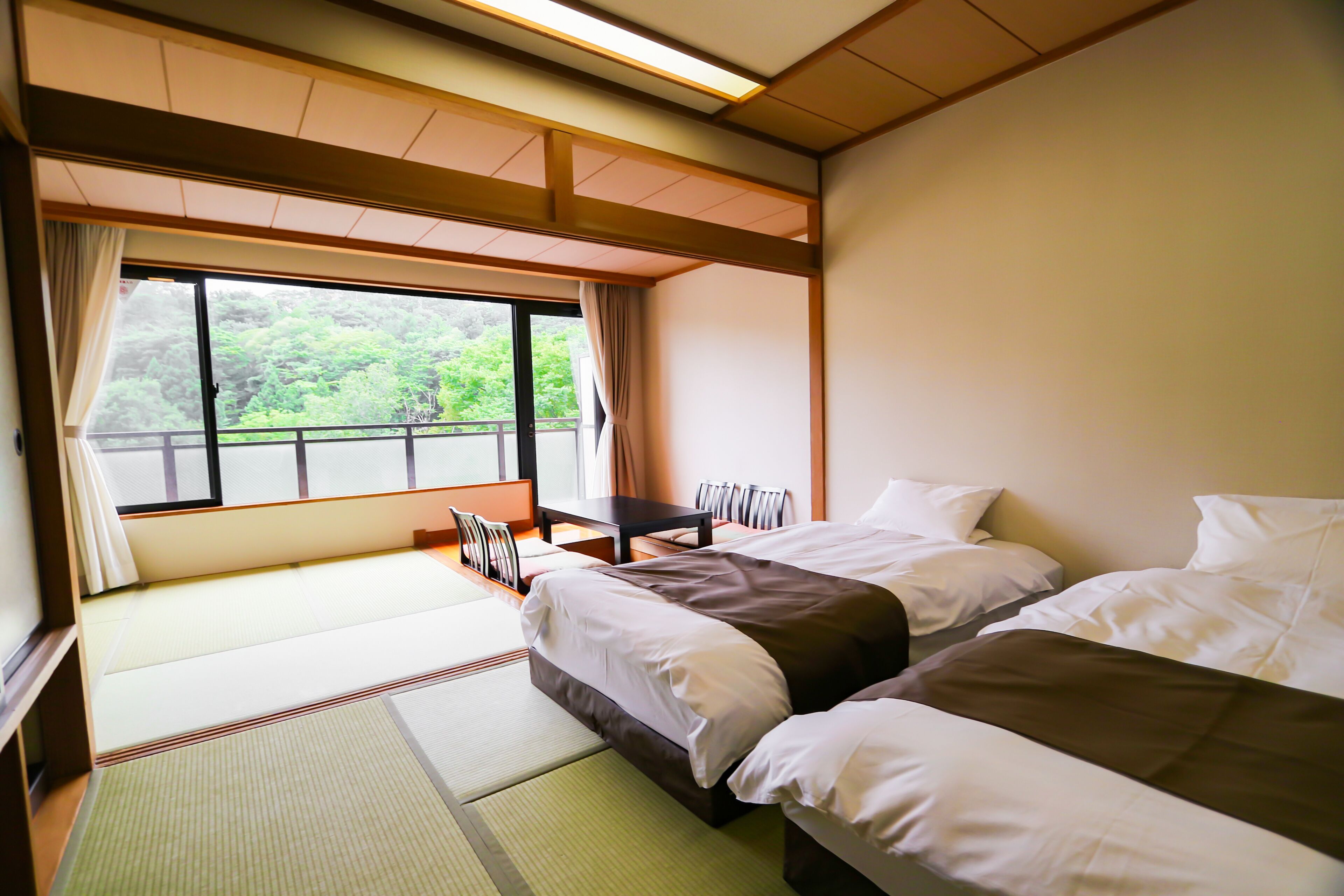 A hotel room with tatami flooring, a large window with a view of greenery, a desk, and two beds.