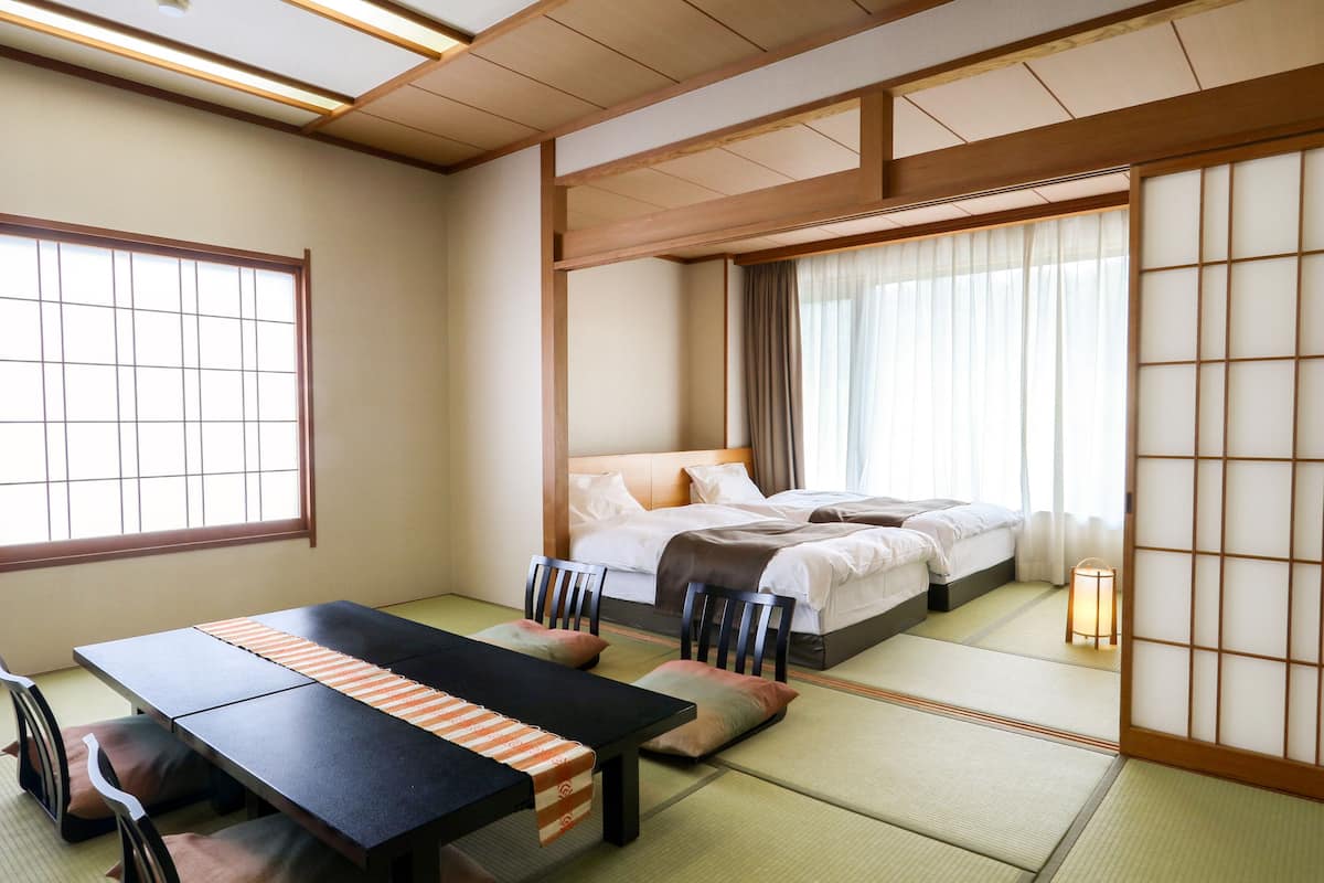 A traditional Japanese-style room with tatami flooring, a low table with cushions, a bed with a wooden headboard, and a sliding shoji screen.