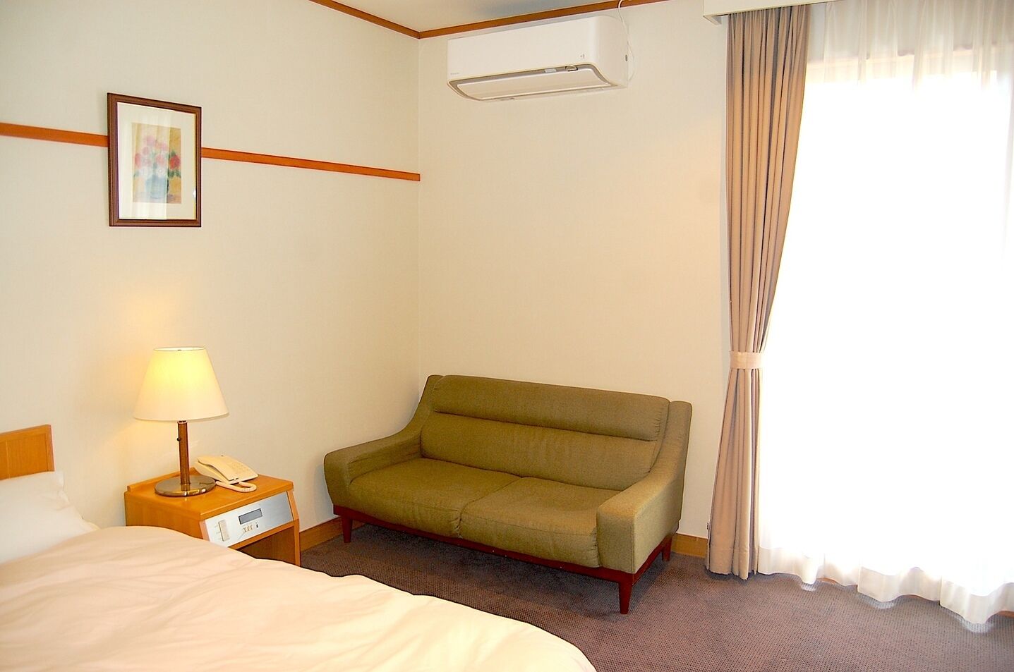Ett hotellrum med en säng, en stol, ett sängbord med en lampa och en telefon, en tavla på väggen och ett fönster med gardiner.