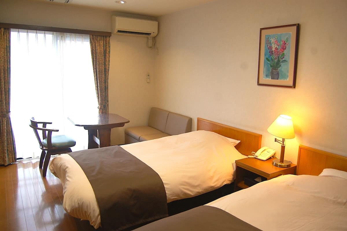 A hotel room with two beds, a desk, a chair, a lamp, and a painting on the wall.