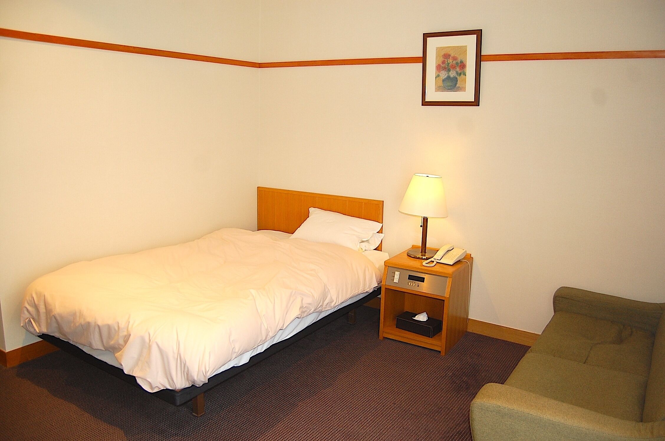 A hotel room with a single bed, a bedside table with a lamp, a chair, and a framed picture on the wall.