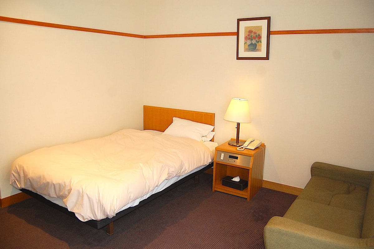 A hotel room with a single bed, a bedside table with a lamp, a chair, and a framed picture on the wall.