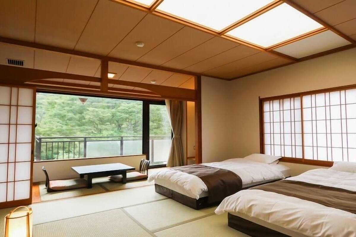 A traditional Japanese-style room with tatami flooring, two beds, a small table, and a view of greenery outside.