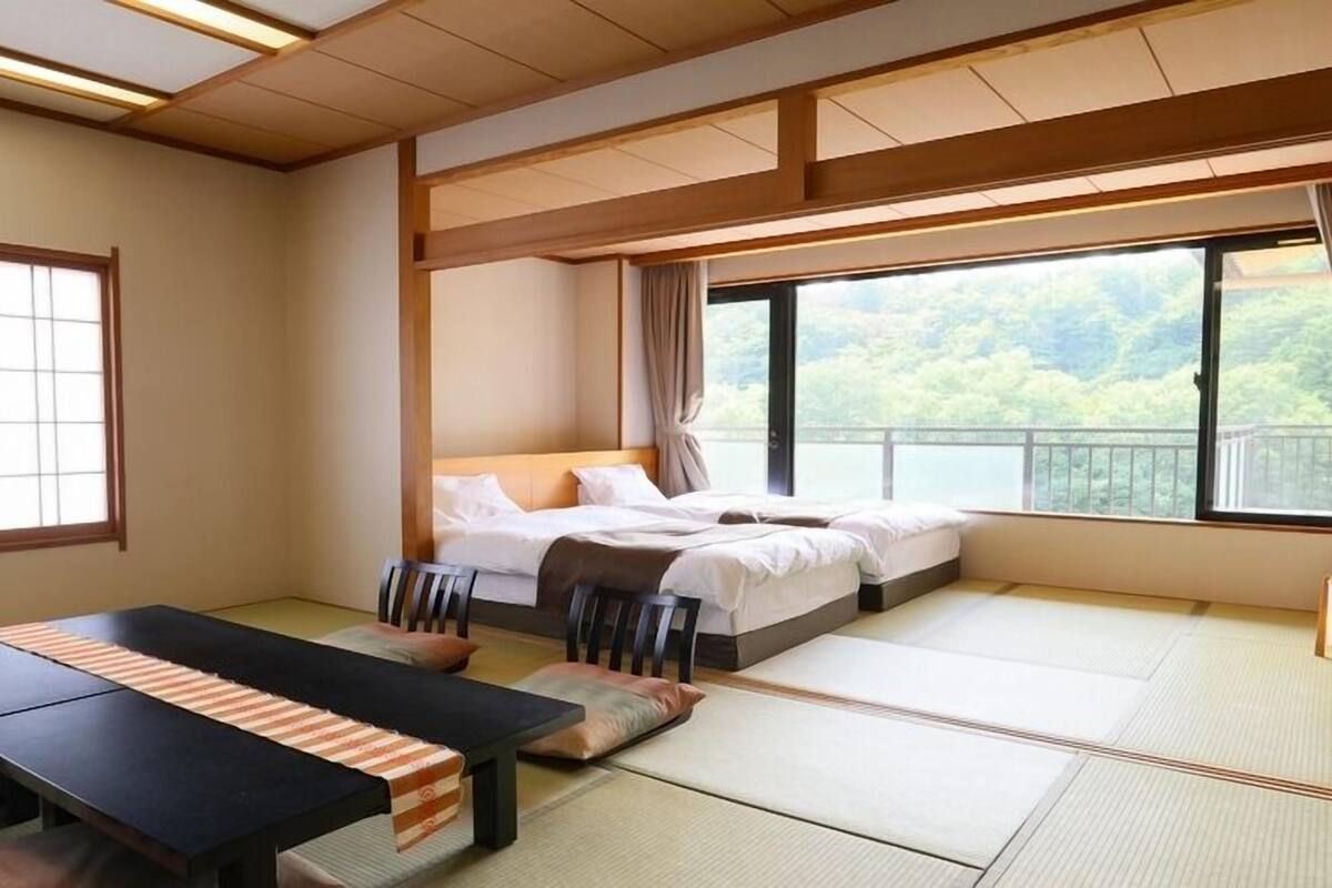 A traditional Japanese-style room with tatami flooring, a low bed, a dining table with cushions, and large windows offering a view of greenery.