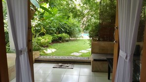 Vista a través de una puerta abierta hacia un jardín con suelo de baldosas, un banco y una pequeña mesa con un felpudo.