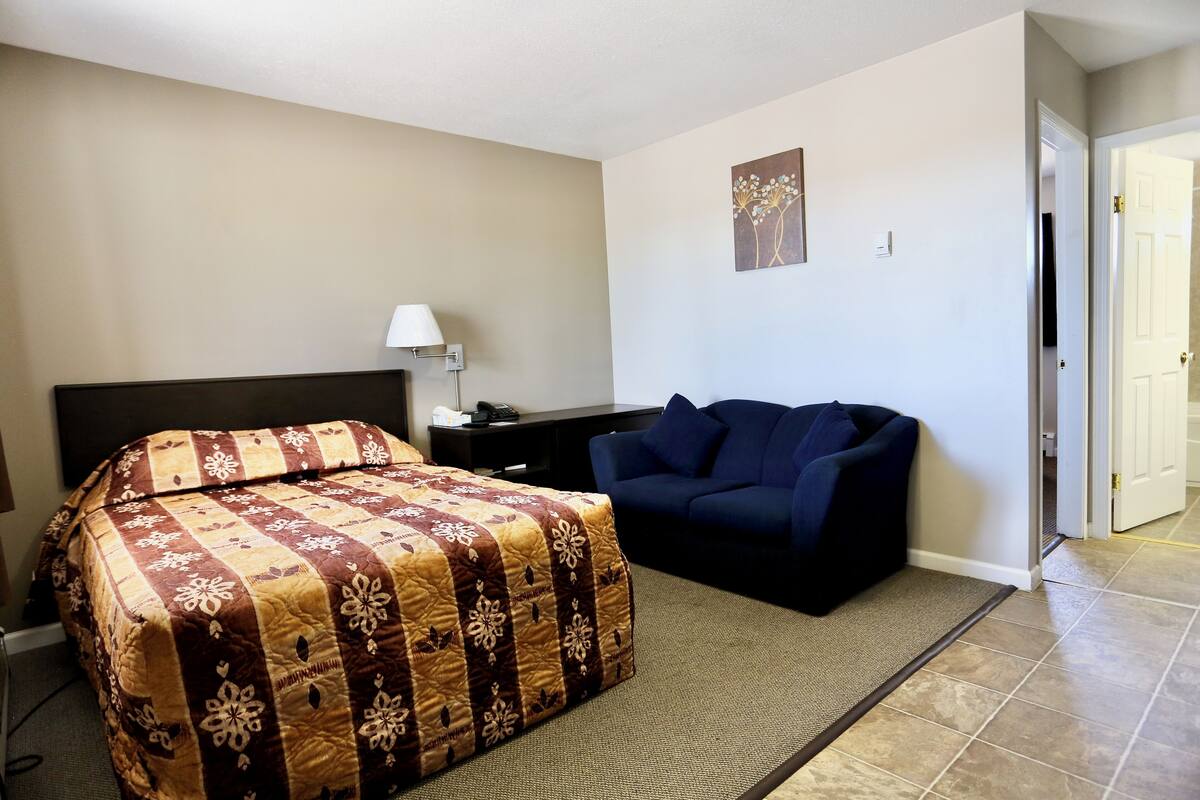 A hotel room with a bed, a sofa, a desk with a lamp, and a door leading to another room.