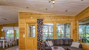 Interior de una cabaña moderna de troncos con piso de madera, un sofá seccional gris, una pequeña mesa de centro de madera y grandes ventanas que ofrecen vistas al exterior.