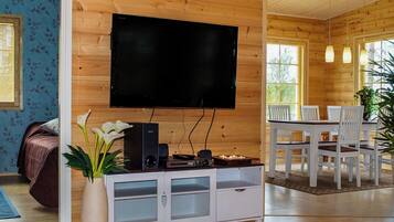 Sala de estar com televisão de tela plana na parede, rack branco com alto-falantes, área de jantar com cadeiras brancas e uma grande janela que permite entrada de luz natural.