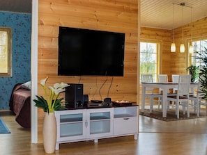 Een woonkamer met een flatscreen tv aan de muur, een witte tv-meubel met speakers, een eethoek met witte stoelen, en een groot raam dat natuurlijk licht binnenlaat.