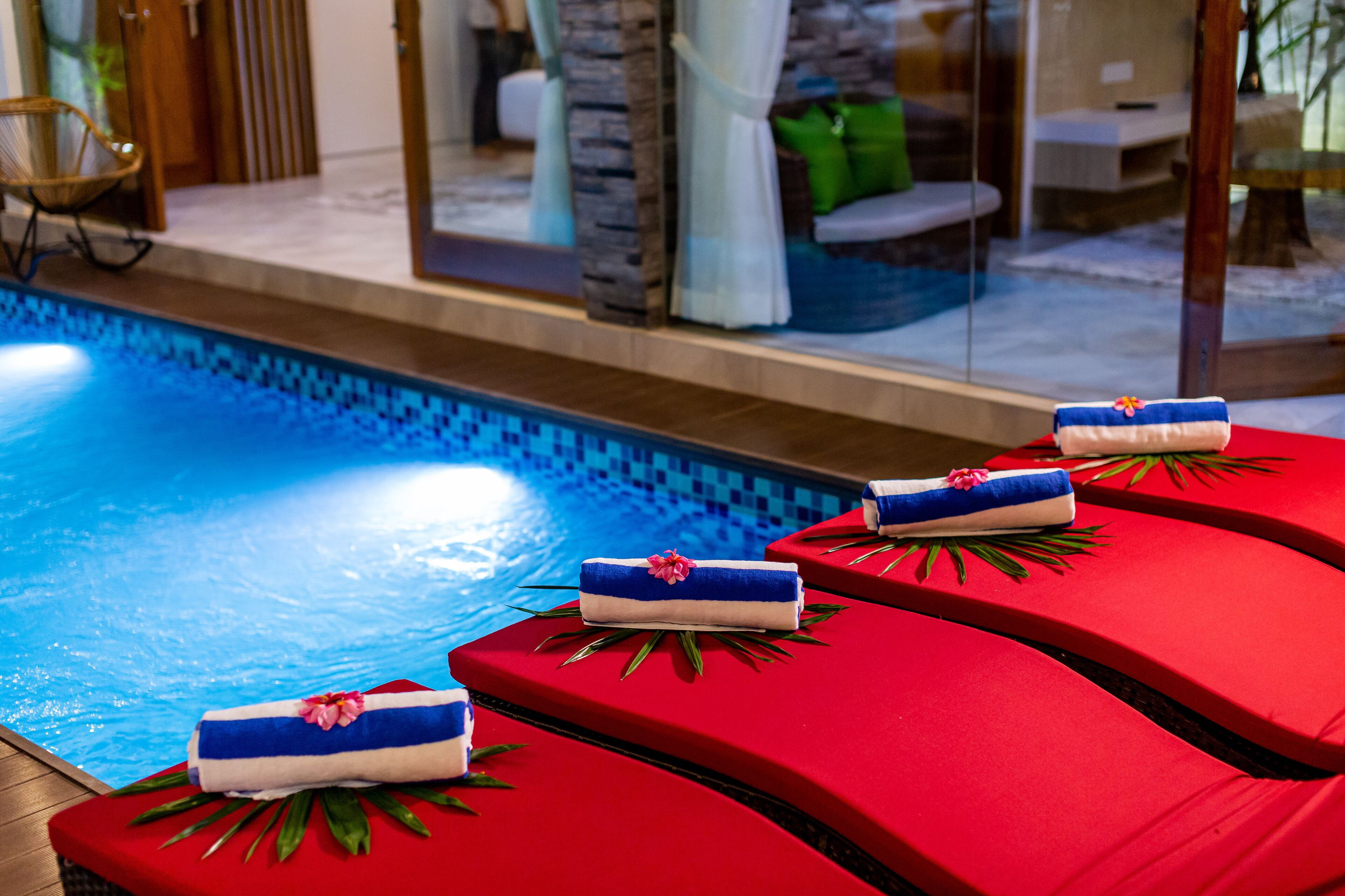 A poolside area with red lounge chairs and towels, a clear blue pool, and a glass wall with a view of the interior.