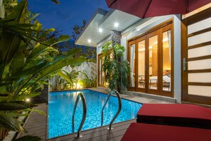 A swimming pool with a wooden deck, surrounded by tropical plants and a modern house with glass doors.