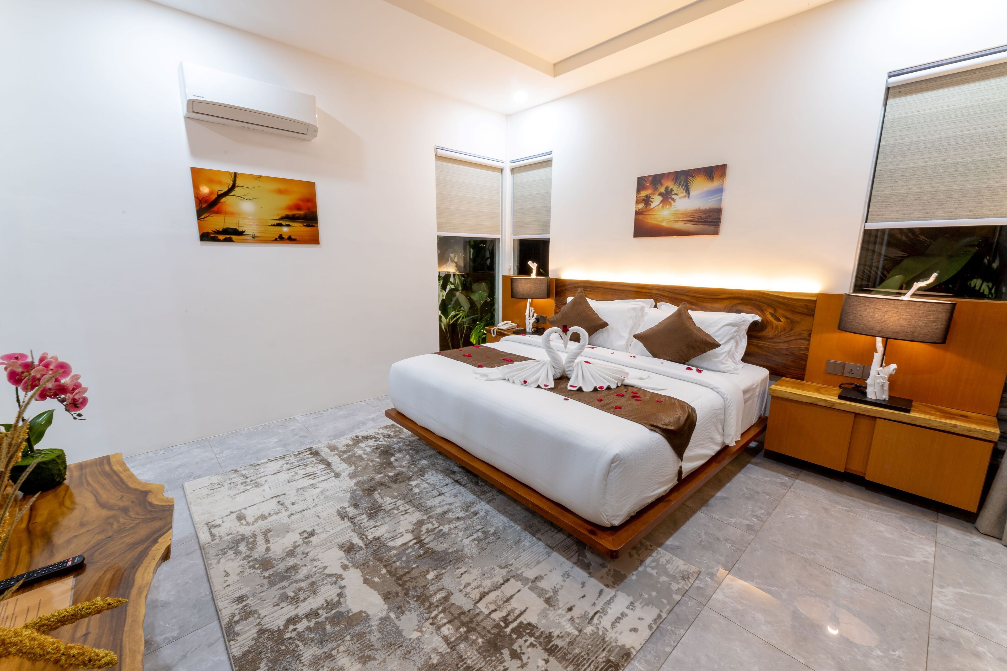 A modern hotel room with a large bed, a wooden bedside table, a nightstand, a desk, and a large window.