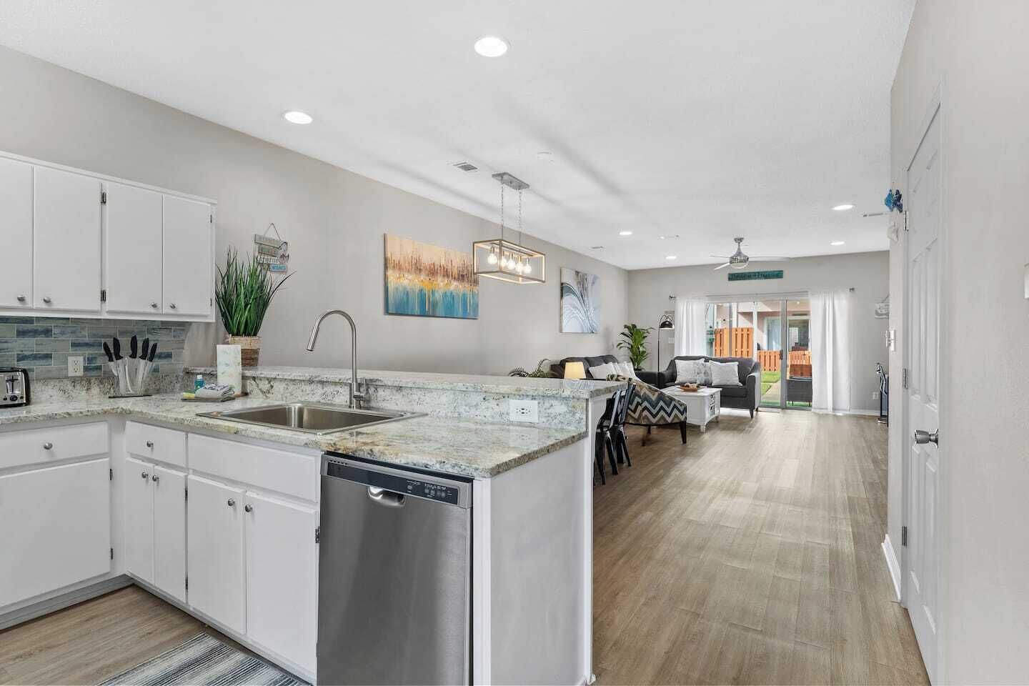 A modern kitchen with white cabinets, a dishwasher, and a granite countertop. There is a dining area with a table and chairs, and a living room with a sofa and a coffee table.