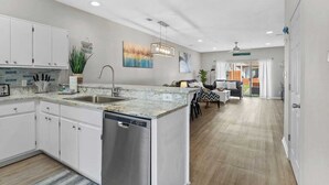 Une cuisine moderne avec des meubles blancs, un lave-vaisselle et un plan de travail en granit. On y trouve un coin repas avec une table et des chaises, ainsi qu’un salon avec un canapé et une table basse.