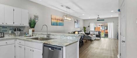A modern kitchen with white cabinets, a dishwasher, and a granite countertop. There is a dining area with a table and chairs, and a living room with a sofa and a coffee table.
