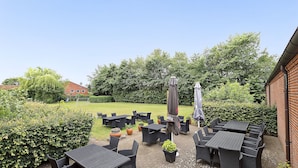 Un espace extérieur aménagé en terrasse, avec plusieurs tables et chaises, entouré de verdure et bordé d’un bâtiment en briques.