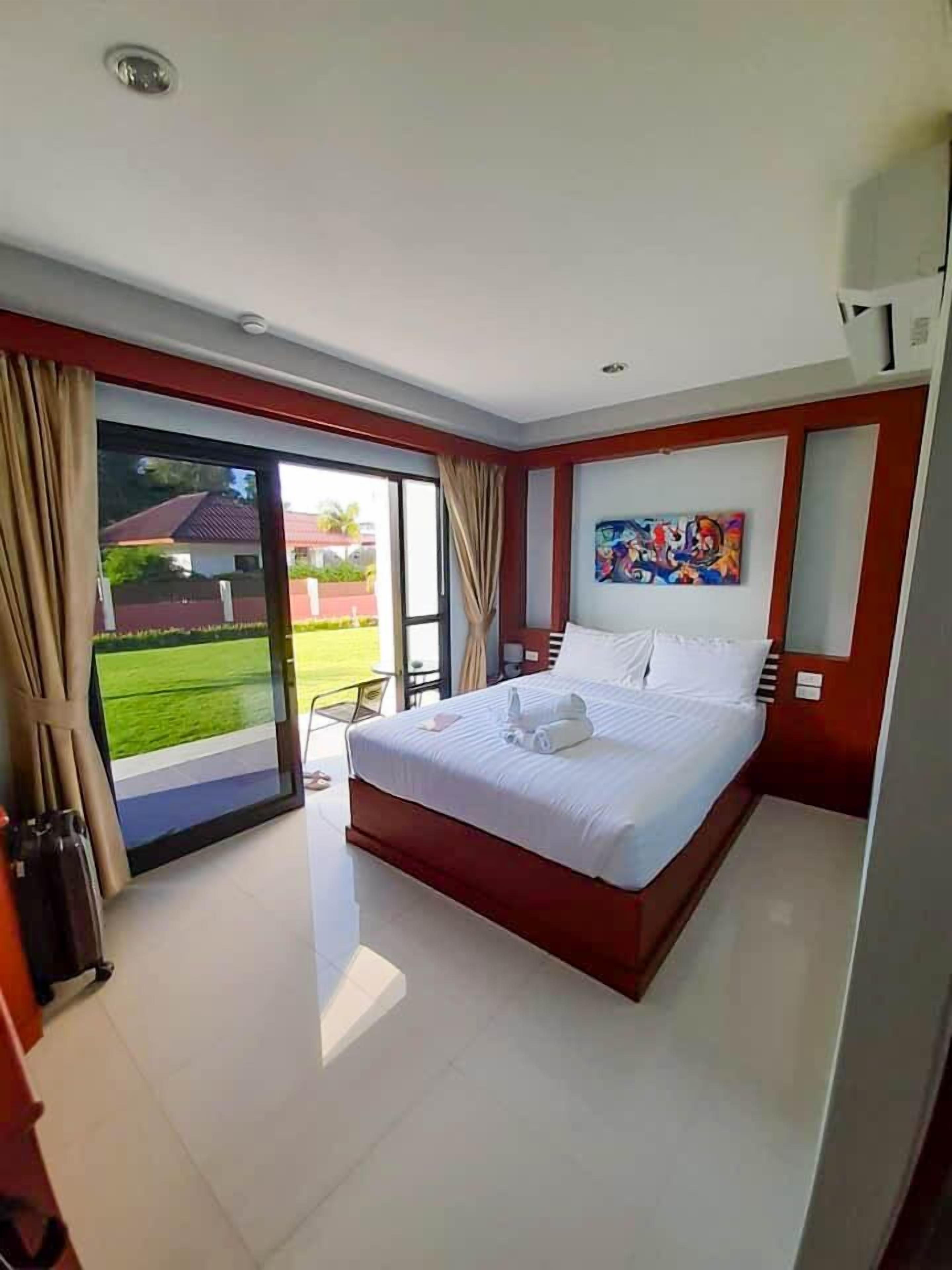 A hotel room with a large bed, a painting on the wall, and a view of a garden through the sliding glass door.