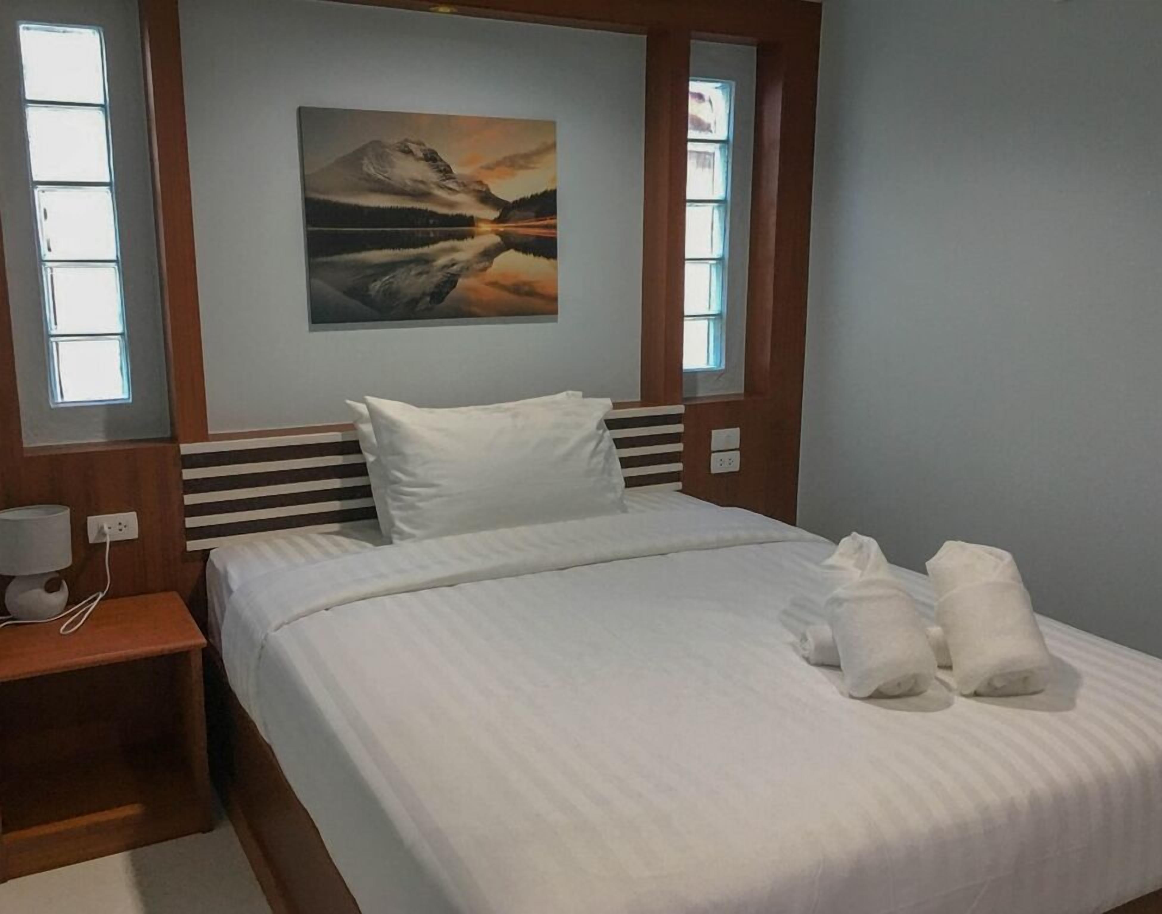 A neatly made bed with white linens and two towels, a wooden headboard, a nightstand with a lamp, and a painting of a mountain landscape on the wall.
