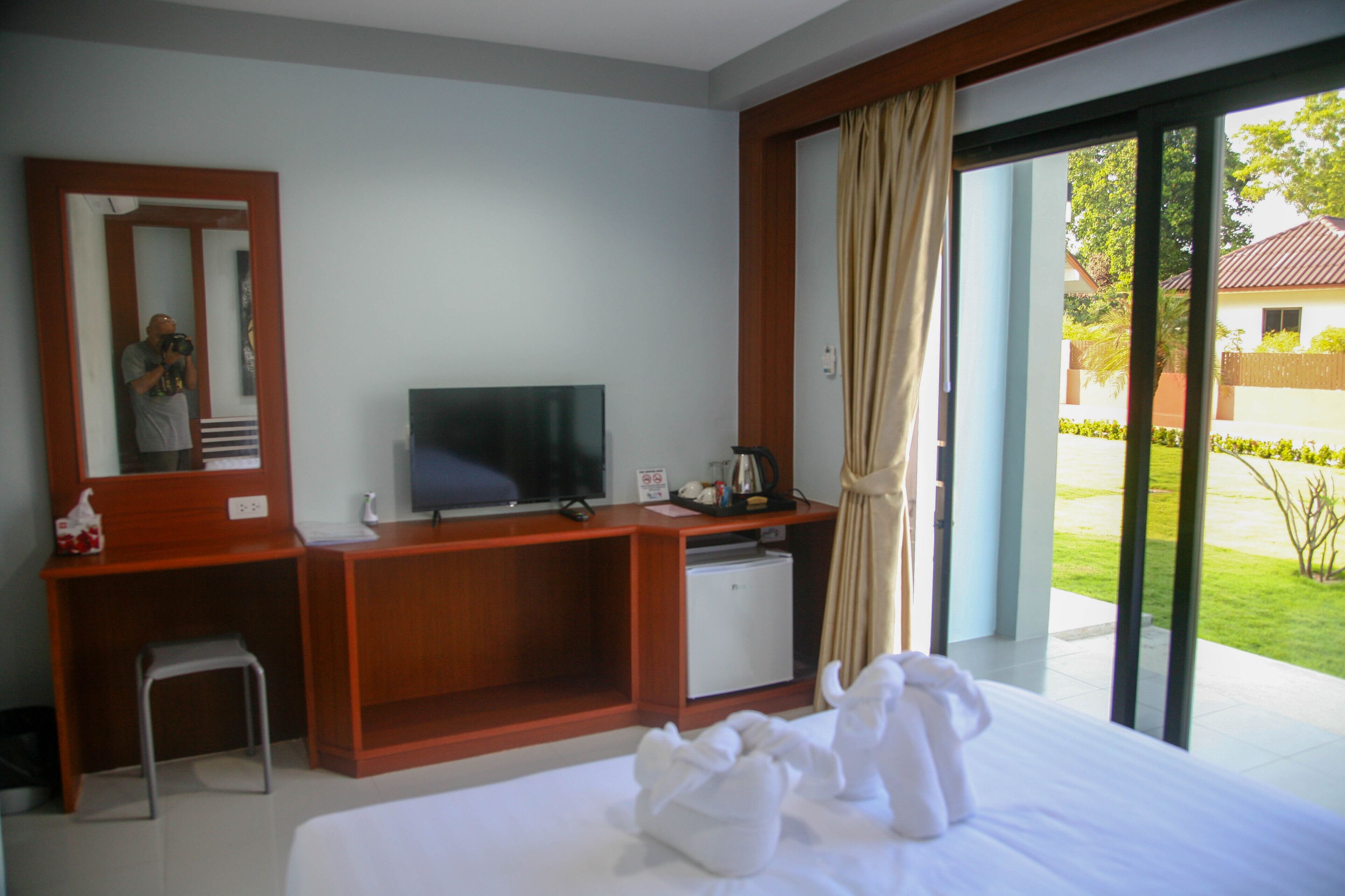 A hotel room with a bed, a desk with a television, a small refrigerator, and a view of a garden through a sliding glass door.
