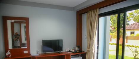 A hotel room with a bed, a desk with a television, a small refrigerator, and a view of a garden through a sliding glass door.