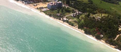 An aerial view of a coastal area with a beach, a large building complex, and surrounding greenery.