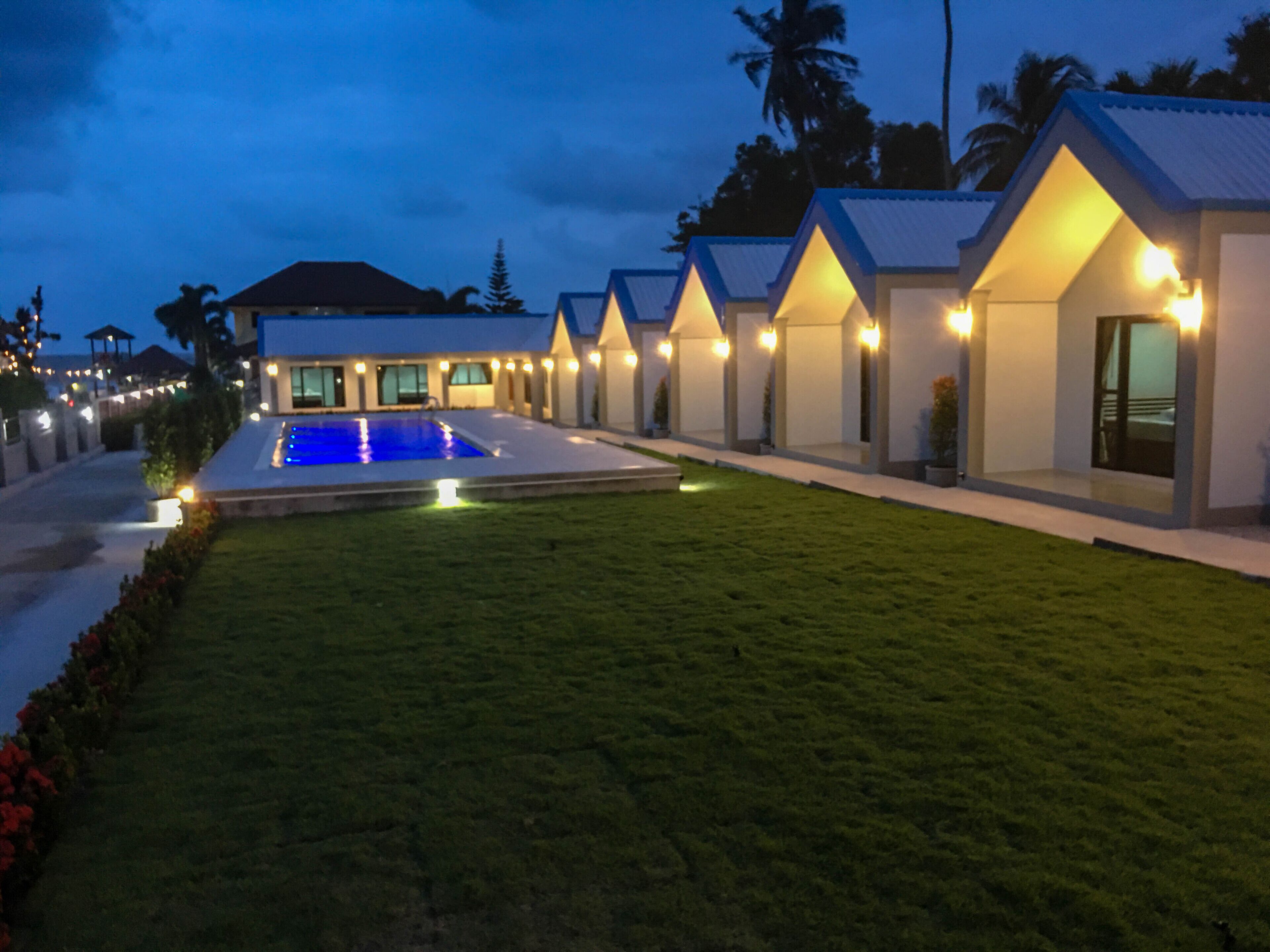 Un resort moderno con piscina, césped impecable y varias villas para huéspedes con iluminación exterior.