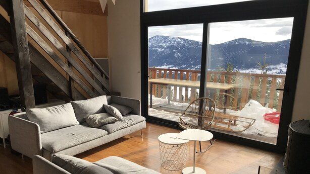 A modern living room with a grey sofa, a small round table, and a chair. A wooden staircase is visible in the background. - Chalet Les Angles 1900m (Les Angles)